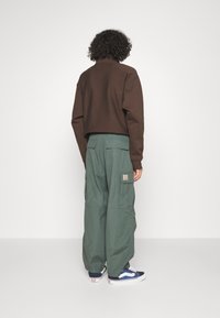 Brązowy krótki sweter, zielone spodnie cargo z bocznymi kieszeniami i niebieskie trampki. Miękki materiał o luźnym kroju z widocznym logo marki.