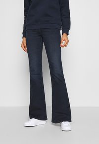 Person som bär mörka utsvängda jeans, mörk sweatshirt och vita sneakers, stående mot en enfärgad ljus bakgrund.