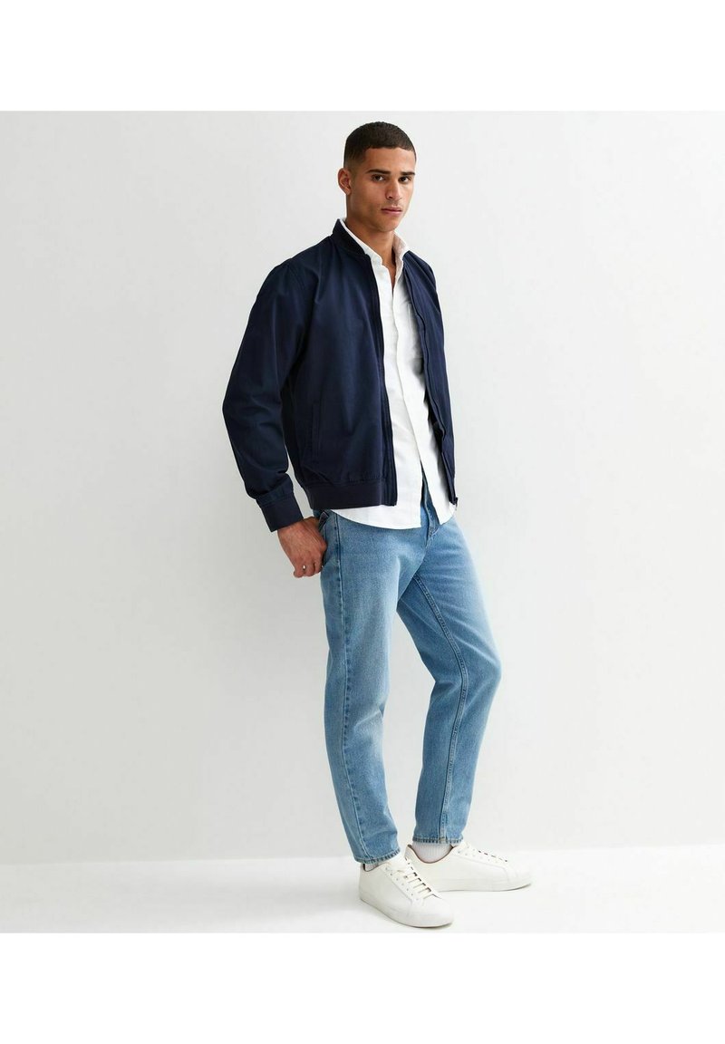Marine bomberjack met een geribbelde kraag, een wit overhemd eronder, lichtblauwe jeans en witte sneakers. Vlotte stof, relaxed fit.