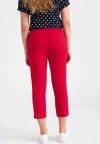 Pantalon rouge raccourci en tissu lisse, avec une coupe ajustée et une taille mi-haute, associé à des chaussures blanches.