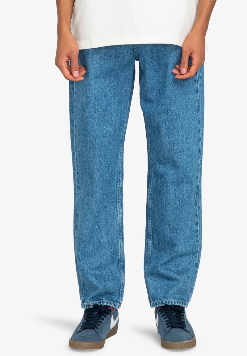 Jeans de mezclilla azul con corte recto, que presentan una textura descolorida y costuras en contraste. Combinados con zapatillas deportivas grises.
