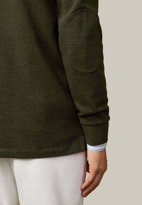 Długa koszula w oliwkowej zieleni z okrągłym kołnierzem i naszywkami na łokciach, wykonana z teksturowanej tkaniny, z warstwowym, niebiesko-paskowanym mankietem pod spodem.