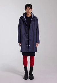 Navy raincoat with a zippered hood, silver snap buttons, and a smooth, water-resistant texture. Worn over a black dress and red tights.