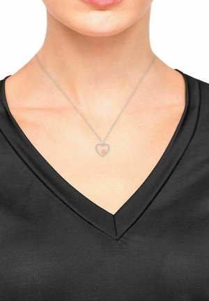 Zilveren ketting met een hartvormige hanger, met een klein innerlijk hartdetail, gedragen met een zwarte V-hals top.