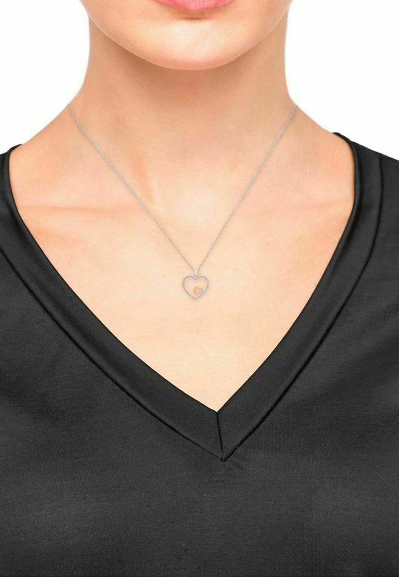 Silberne Kettenhalskette mit einem herzförmigen Anhänger, der ein kleines inneres Herzdetail aufweist, getragen mit einem schwarzen V-Ausschnitt-Oberteil.