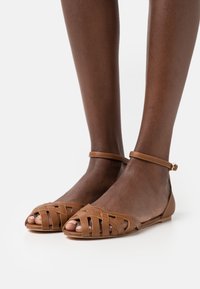 Bruna lädersandaler har en öppen tåsdesign med korsade remmar och en smal ankelrem, platta sulor och en mjuk yta.