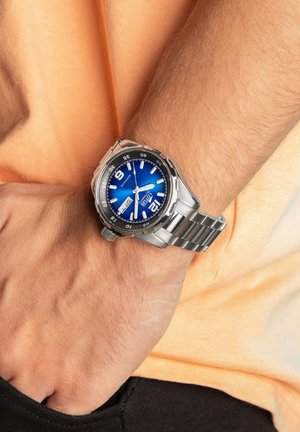 Montre en acier inoxydable argenté avec un cadran bleu, de grands index horaires blancs et un bracelet en maillons métalliques, portée au poignet sur un fond orange.