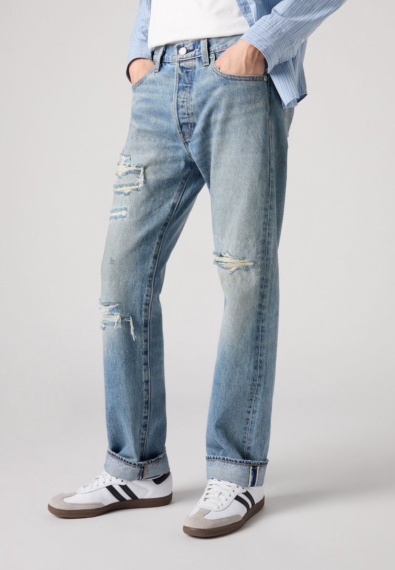 Homme portant un jean bleu clair déchiré, des baskets blanches avec des bandes noires et une chemise rayée bleue, les mains dans les poches.