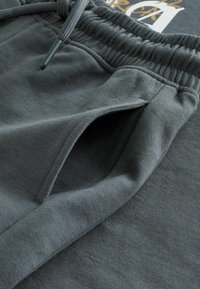 Pantalon de survêtement gris foncé présentant une taille élastique, un cordon en tissu et une poche avant inclinée avec ouverture visible.