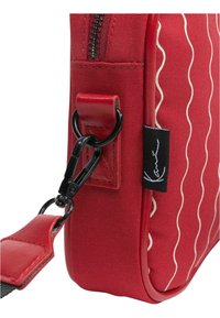 Červená látková taška s vlnkovým vzorem, koženými detaily, zipovým uzávěrem a klipovým popruhem. Na boku je viditelný štítek.