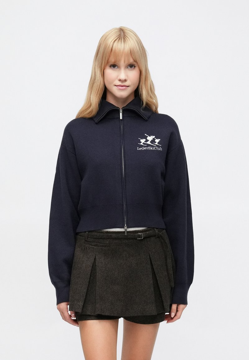 Navy krāsas īsa džemperis ar augstu apkakli, rāvējslēdzēju un izšūtu "LeGetSkiClub" logotipu. Kombinēts ar struktūr�ētu brūnu plisirētu svārku.