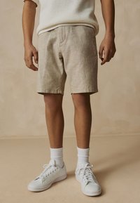 Beige linen shorts and white sneakers with textured fabric, paired with white ribbed socks, standing against a neutral background.