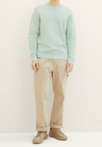 Person in einem hellgrünen Langarmpullover, beigen hochgekrempelten Hosen mit aufgesetzten Taschen und beigen High-Top-Sneakern, der auf einem weißen Boden steht.