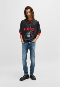Maglietta nera a maniche corte con dettagli rossi, caratterizzata da un design grafico di un ragno e il testo "HUGO 93". Indossata con jeans azzurri chiari e scarpe nere.