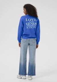 Femme aux cheveux bouclés portant un sweat-shirt bleu avec le texte "LOVE GENERATION" dans le dos, un jean clair délavé et des baskets blanches, se tenant de dos.