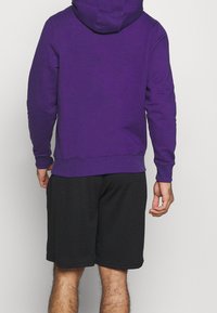 Sudadera con capucha morada, con puños y dobladillo de canalé, combinada con pantalones cortos negros texturizados. Vista trasera que muestra detalles de ajuste y tela.