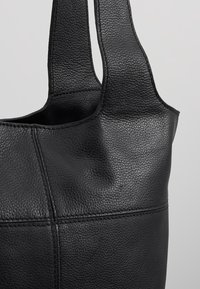 Sac à main en cuir texturé noir avec larges anses et coutures apparentes sur un fond gris clair.