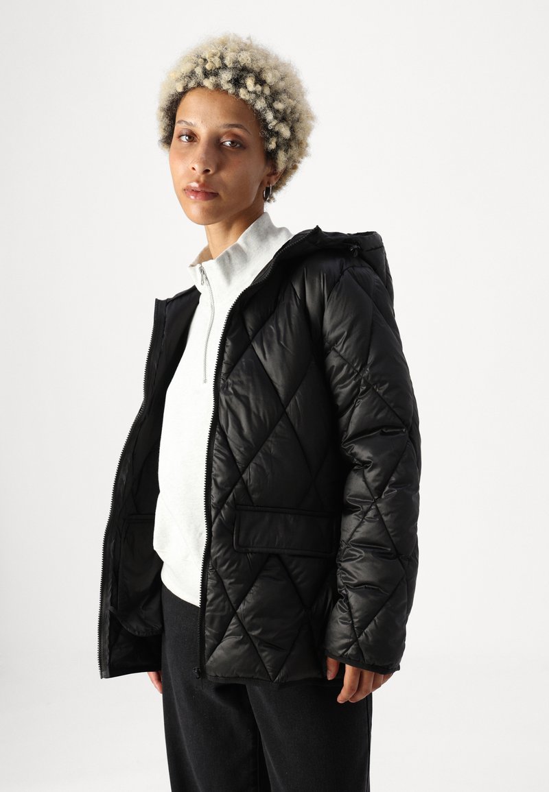 Chaqueta acolchada negra con capucha, que presenta un cierre de cremallera, bolsillos y una textura suave. Llevada sobre una blusa gris sin cuello.