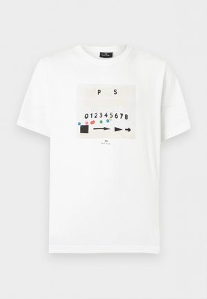 Ung mand med krøllet hår iført en hvid grafisk t-shirt, lyseblå jeans med vide ben og hvide sneakers, stående mod en ensfarvet baggrund.