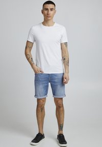 Hvit t-skjorte med korte ermer kombinert med lys blå denimshorts med oppbrett og svarte sneakers. Bakgrunnen er enkel og nøytral.