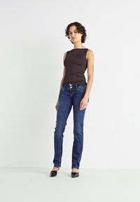 Top sin mangas marrón combinado con jeans de denim ajustados en azul oscuro, con tres botones en la cintura y bailarinas negras. Fondo liso.