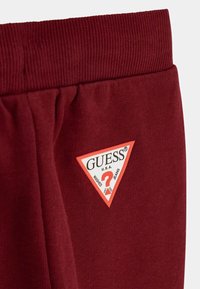 Burgundy-huonefarkut, joissa on ribbikuvioitu vyötärö ja etuosassa kolmikulmainen punavalkoinen logopatch, jossa lukee "GUESS U.S.A."