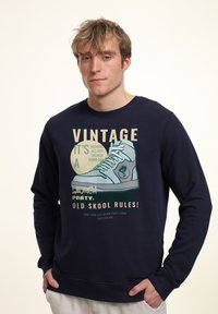 Felpa navy con grafica di una sneaker e testo. Tessuto morbido, collo a giro e maniche lunghe. Il design frontale include una tipografia giocosa.