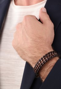 Pulsera de cuero trenzado marrón con acentos de cobre, con un diseño plano que se envuelve alrededor de la muñeca, parcialmente oculta por una mano.