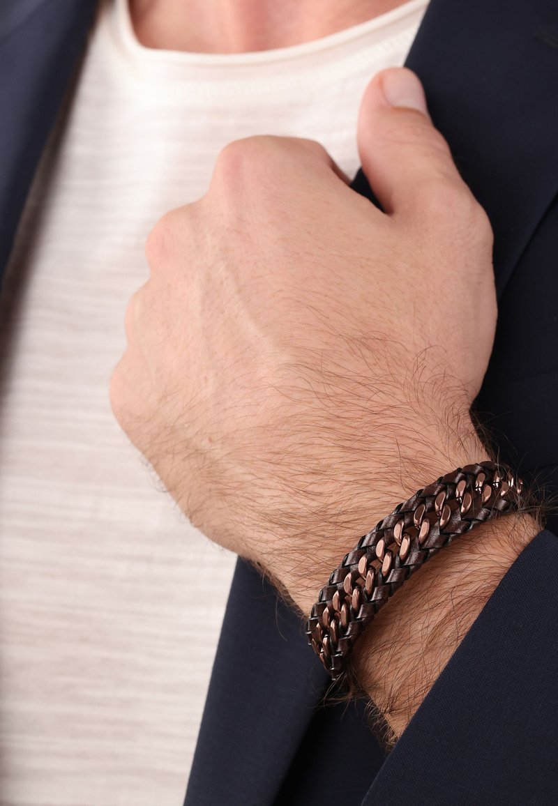 Pulsera de cuero trenzado marrón con acentos de cobre, con un diseño plano que se envuelve alrededor de la muñeca, parcialmente oculta por una mano.