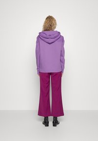 Sudadera con capucha morada de corte relajado, combinada con pantalones burgundy de pierna ancha y botas negras de tobillo, tomada desde atrás.