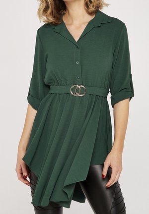 Femme portant une tunique asymétrique vert foncé ceinturée avec des manches retroussées, sur des leggings noirs texturés.