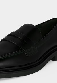Mocassin en cuir noir avec un petit talon et une bride décorative sur le dessus, vu de côté sur un fond blanc.