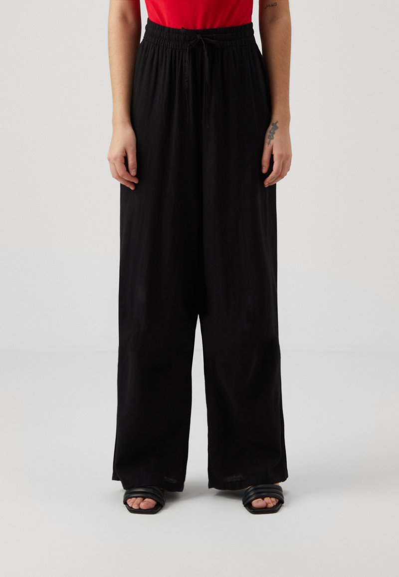 Pantalons larges noirs avec une taille à cordon, fabriqués en tissu léger. Associés à un haut rouge et des sandales noires.