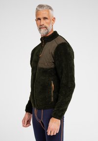 Grøn fleecejakke med brune detaljer og lynlås foran. Har en brystlomme, sidelommer og tekstureret stof for varme.