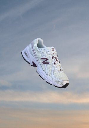 Бял и лилав маратонка New Balance, висящ на фона на облачно небе.
