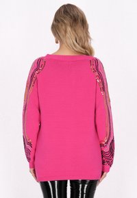 Maglione fucsia con maniche lunghe, caratterizzato da motivi di paillettes ondulati e multicolori lungo le spalle. La modella indossa pantaloni neri in vernice.