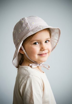 Young girl smiling, wearing a light pink sun hat with white daisies and a matching tied chin strap, beige shirt, against plain gray background.