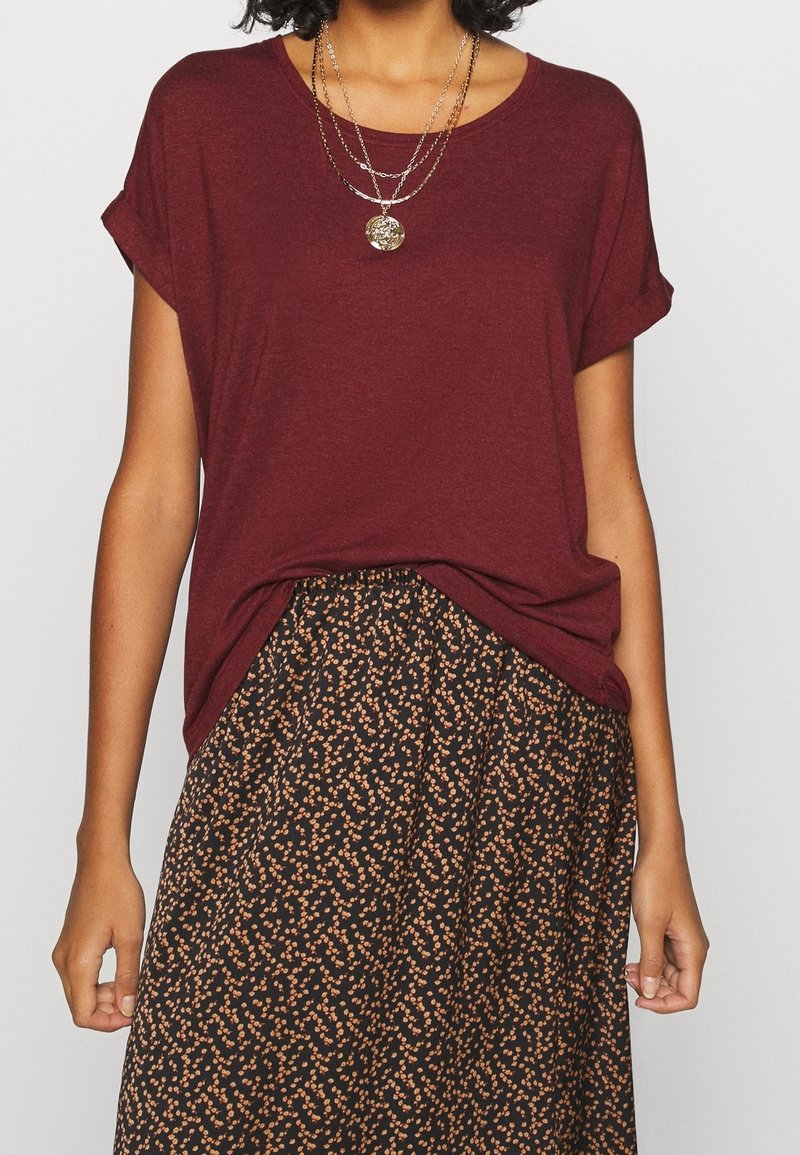 Femme portant un chemisier bordeaux à manches courtes rentré dans une jupe noire à motifs floraux orange et des colliers en or superposés.