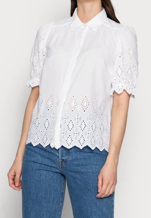 Vrouw draagt een witte blouse met korte mouwen, opengewerkte details en geschulpte randen, gecombineerd met blauwe spijkerbroek.