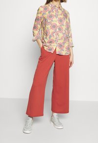 Camisa con estampado floral en amarillo, rosa y negro con diseño de botones. Combinada con pantalones anchos rojos y zapatillas gris claro.
