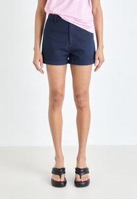 Kvinna iförd mörkblå shorts med rullad fåll, överdel med rosa och vita ränder, samt svarta vadderade plattformssandaler, stående mot en enfärgad vit bakgrund.