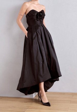 Czarna, asymetryczna sukienka bez ramiączek z kwiatowym akcentem na biuście, z kieszeniami, zestawiona z czarnymi, spiczastymi szpilkami, na drewnianej podłodze, na tle jednolitej ściany.