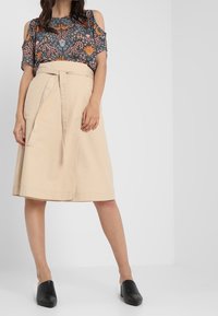 Dunkelbluse mit Blumenmuster und kurzen Ärmeln, kombiniert mit einem beigefarbenen, taillierten, knielangen Rock. Die Schuhe sind schwarze, geschlossene Slippers.