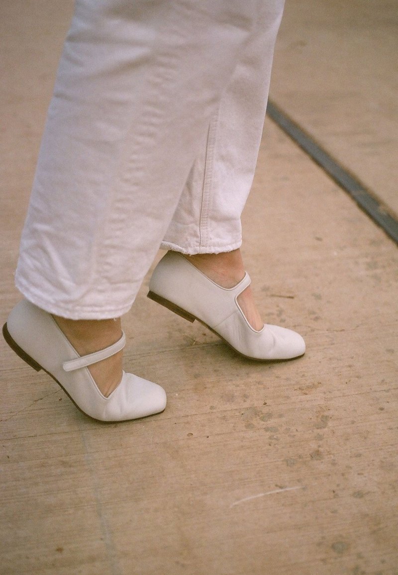 Zapatos Mary Jane de cuero blanco con punta cuadrada y corte asimétrico, que cuentan con una correa delgada sobre el empeine y un bajo tacón marrón.