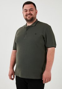 Tmavo zelená polo košela z mäkkej, štruktúrovanej tkaniny, s trojitém gombíkovým zapínaním a decentným logom na ľavej hrudi. Krátke rukávy a rovný lem.