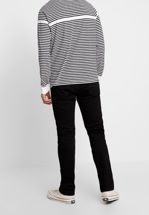 Osoba stojąca, ubrana w czarne spodnie, białe trampki i koszulę z długim rękawem w czarno-białe poziome paski, stojąca tyłem do kamery.