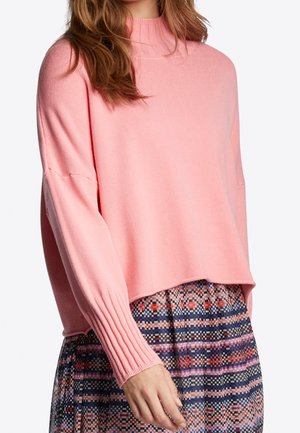 Kobieta ubrana w luźny różowy sweter z prążkowanymi mankietami i kołnierzem, zestawiony z wielokolorową, wzorzystą spódnicą, widoczna od szyi do bioder.