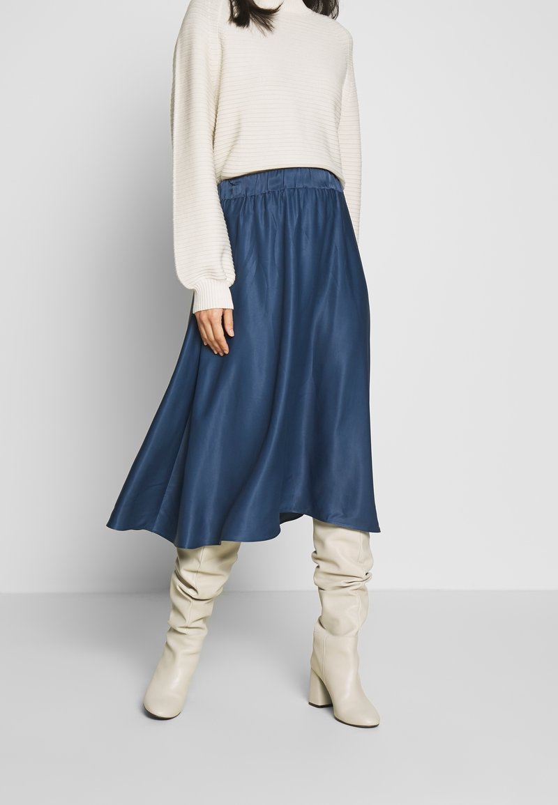Femme portant un pull crème côtelé, une jupe midi bleu marine et des bottes à talons blanches décontractées devant un fond uni gris clair.
