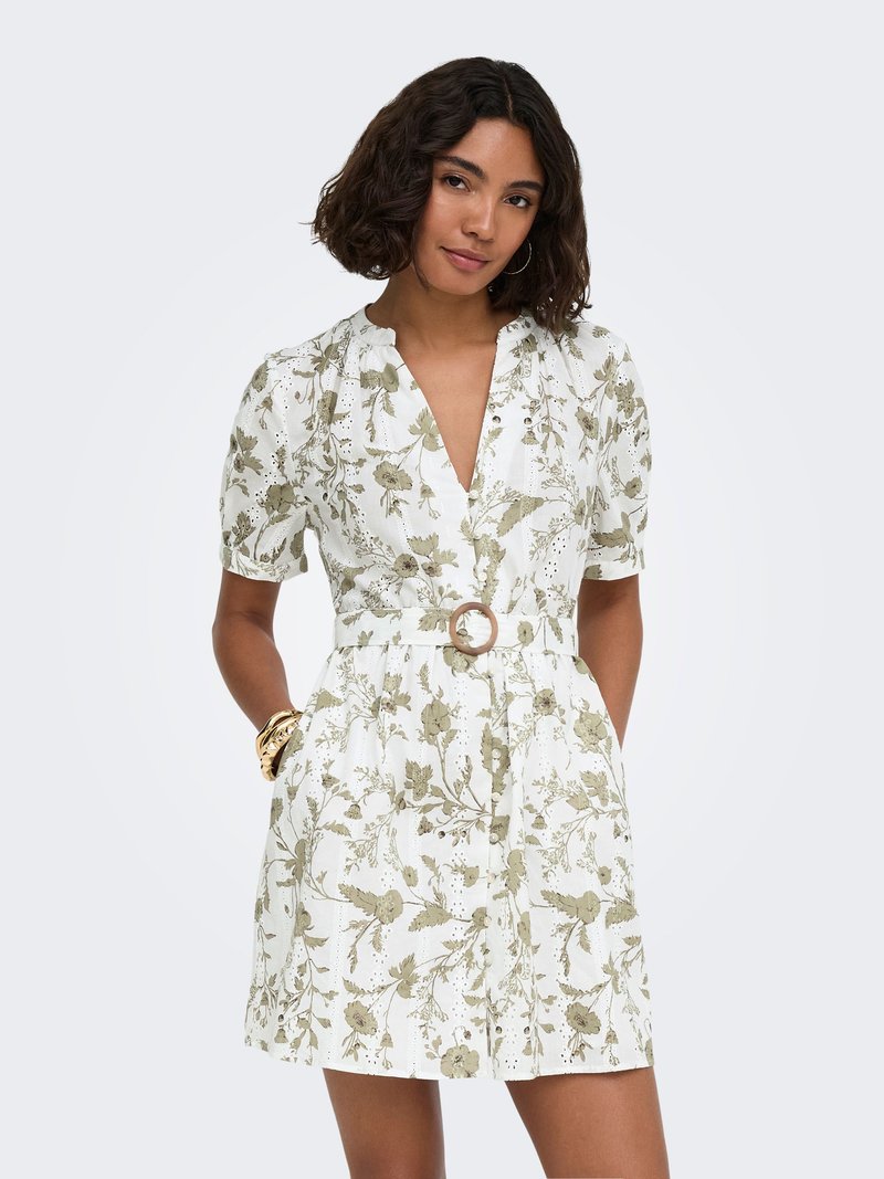 Femme aux cheveux bouclés mi-longs portant une robe blanche à fleurs à manches courtes avec une ceinture à la taille et les mains dans les poches, sur un fond uni.