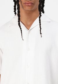 White short-sleeve shirt made of textured fabric with subtle vertical stripes, featuring a notched collar and button closure.